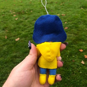 May include: A small felt doll wearing a yellow raincoat and blue hat. The doll is holding a small white object in its hand.