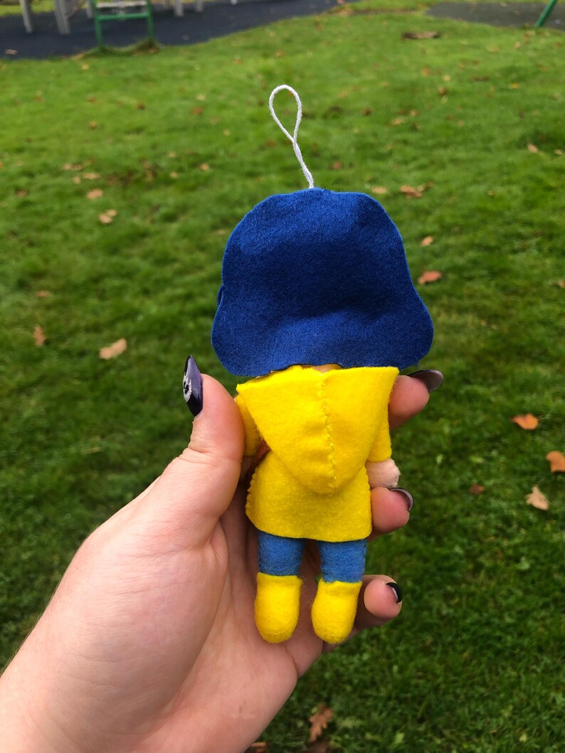 May include: A small felt doll wearing a yellow raincoat and blue hat. The doll is holding a small white object in its hand.