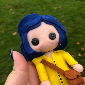 May include: A felt doll with blue hair, a yellow raincoat, and a brown bag. The doll has black button eyes and a light brown face.
