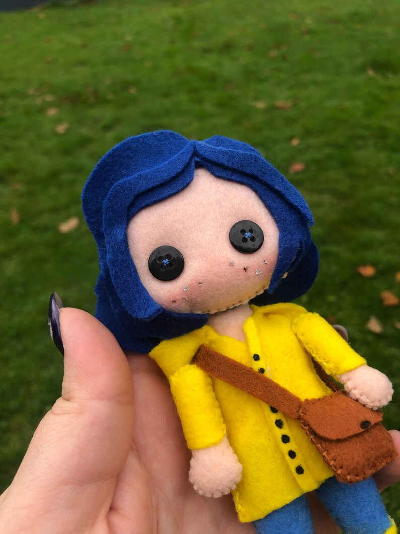 May include: A felt doll with blue hair, a yellow raincoat, and a brown bag. The doll has black button eyes and a light brown face.