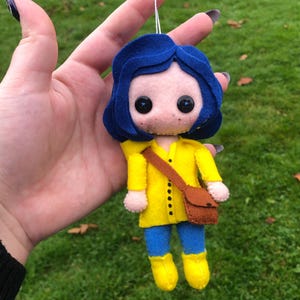 May include: A felt doll of a girl with blue hair, wearing a yellow raincoat, blue pants, and brown boots. She is holding a brown bag.