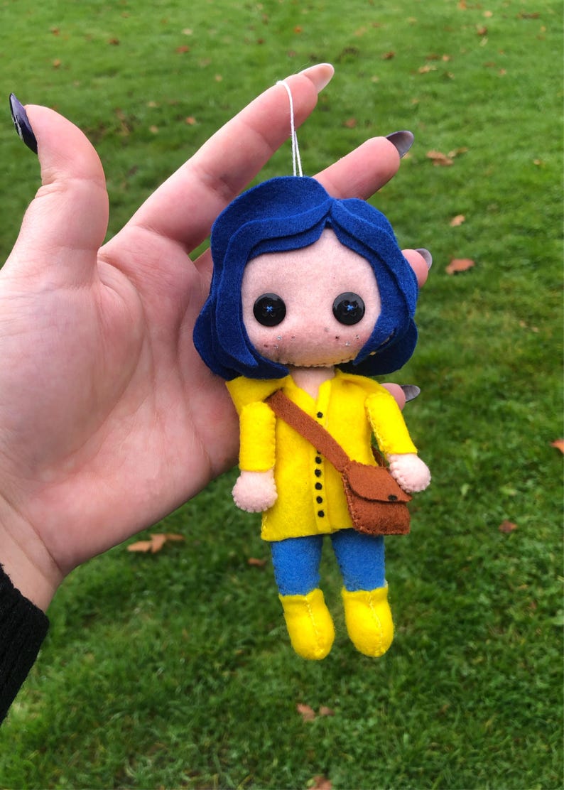 May include: A felt doll of a girl with blue hair, wearing a yellow raincoat, blue pants, and brown boots. She is holding a brown bag.