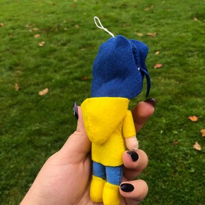 May include: A small felt doll wearing a yellow raincoat and a blue hooded jacket. The doll is holding a small white string in its hand.