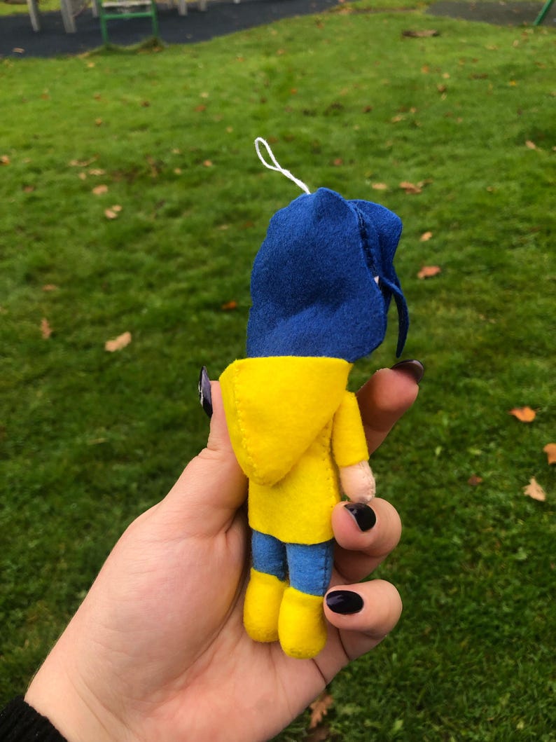 May include: A small felt doll wearing a yellow raincoat and a blue hooded jacket. The doll is holding a small white string in its hand.