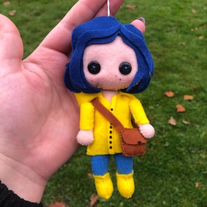 May include: A felt doll of a girl with blue hair wearing a yellow raincoat and brown boots. She is carrying a brown bag.