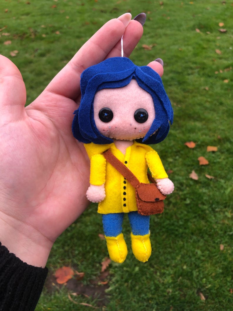 May include: A felt doll of a girl with blue hair wearing a yellow raincoat and brown boots. She is carrying a brown bag.