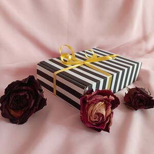 May include: A black and white striped gift box with a yellow ribbon tied in a bow. The box is surrounded by three dried roses on a pink background.