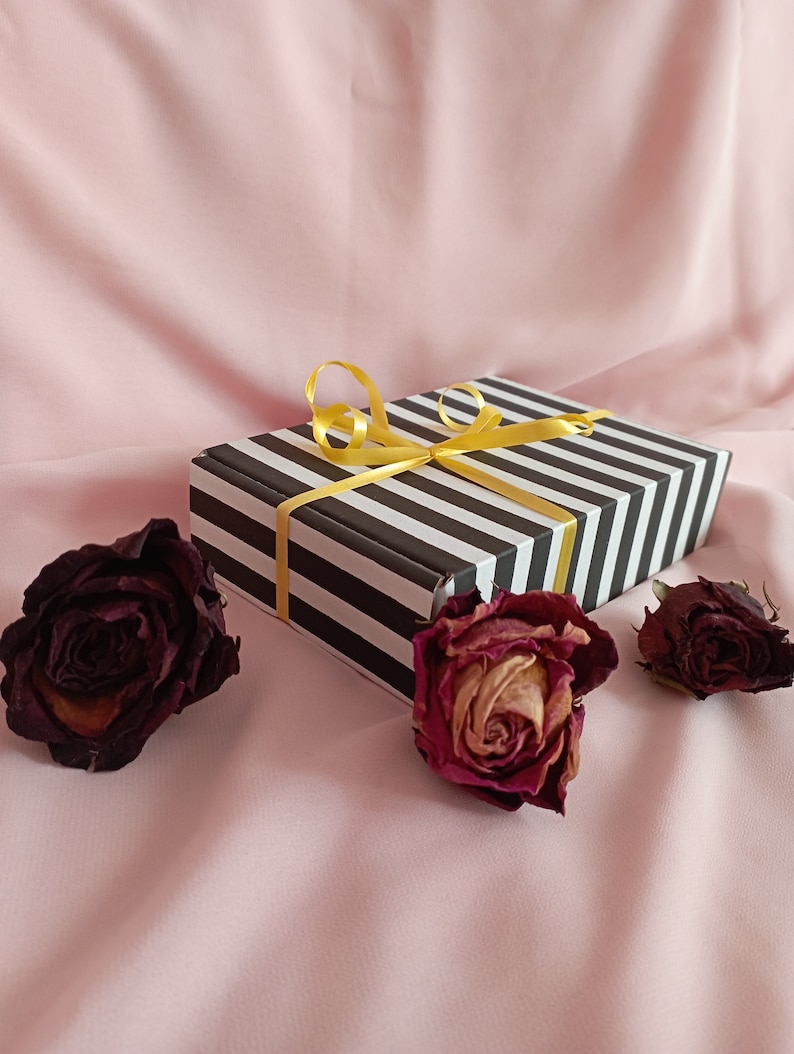 May include: A black and white striped gift box with a yellow ribbon tied in a bow. The box is surrounded by three dried roses on a pink background.