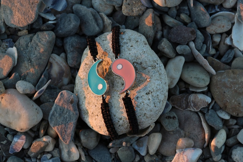 May include: Two black string bracelets with yin yang charms. The charms are silver with blue and pink enamel. The bracelets are on a rock background.