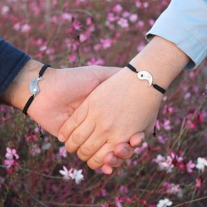 May include: Two people holding hands, each wearing a black string bracelet with a silver yin yang charm.