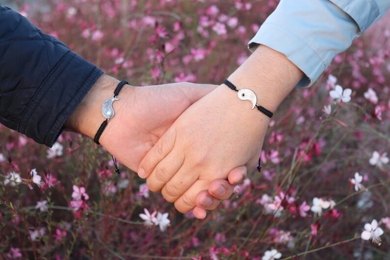 May include: Two people holding hands, each wearing a black string bracelet with a silver yin yang charm.