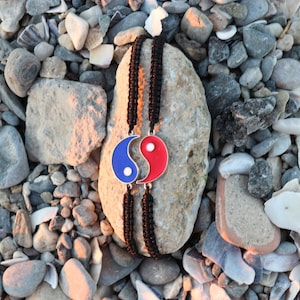 May include: A black cord bracelet with a yin yang charm. The charm is blue and red with a white swirl in the center. The bracelet is on a gray rock with small stones around it.