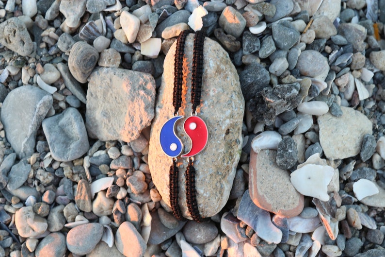 May include: A black cord bracelet with a yin yang charm. The charm is blue and red with a white swirl in the center. The bracelet is on a gray rock with small stones around it.