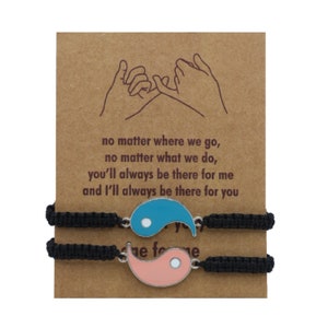 May include: Two black friendship bracelets with a yin yang charm. The charm is made of metal and has a blue and pink enamel finish. The card behind the bracelets reads: "no matter where we go, no matter what we do, you'll always be there for me and I'll always be there for you".