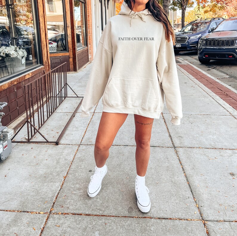 Op de afbeelding: Een licht beige hoodie met een capuchon met trekkoord en een kangoeroezak. De hoodie heeft de tekst "FAITH OVER FEAR" in het zwart op de voorkant gedrukt.