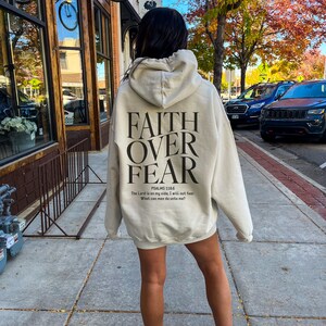 Op de afbeelding: Een beige hoodie met de tekst "FAITH OVER FEAR" en het bijbelvers "PSALMS 118:6 The Lord is on my side, I will not fear. What can man do unto me?" gedrukt op de achterkant.