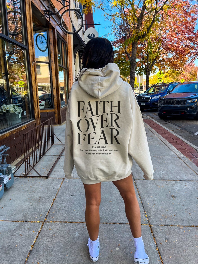 Op de afbeelding: Een beige hoodie met de tekst "FAITH OVER FEAR" en het bijbelvers "PSALMS 118:6 The Lord is on my side, I will not fear. What can man do unto me?" gedrukt op de achterkant.