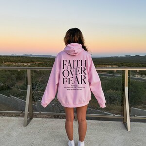 Op de afbeelding: Een roze hoodie met de tekst "FAITH OVER FEAR" en een bijbelvers gedrukt op de achterkant. De hoodie wordt gedragen door een persoon die op een balkon staat met uitzicht op een woestijnlandschap.