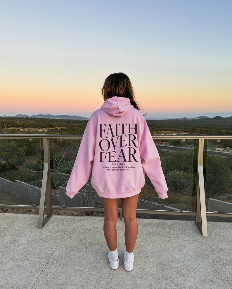 Op de afbeelding: Een roze hoodie met de tekst "FAITH OVER FEAR" en een bijbelvers gedrukt op de achterkant. De hoodie wordt gedragen door een persoon die op een balkon staat met uitzicht op een woestijnlandschap.