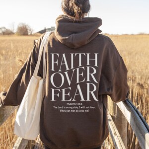 Op de afbeelding: Een bruine hoodie met de woorden "FAITH OVER FEAR" gedrukt op de achterkant. De hoodie bevat ook een Bijbelvers uit Psalm 118:6, "De HEERE is mijn helper, ik zal niet vrezen; wat kan de mens mij doen?"
