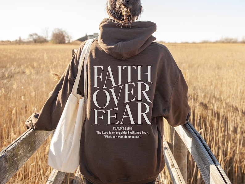 Op de afbeelding: Een bruine hoodie met de woorden "FAITH OVER FEAR" gedrukt op de achterkant. De hoodie bevat ook een Bijbelvers uit Psalm 118:6, "De HEERE is mijn helper, ik zal niet vrezen; wat kan de mens mij doen?"
