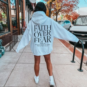 Op de afbeelding: Witte hoodie met de tekst "FAITH OVER FEAR" en een bijbelvers gedrukt op de achterkant.