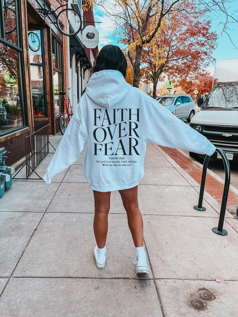 Op de afbeelding: Witte hoodie met de tekst "FAITH OVER FEAR" en een bijbelvers gedrukt op de achterkant.