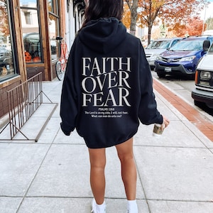 Op de afbeelding: Een zwarte hoodie met de woorden "FAITH OVER FEAR" gedrukt op de achterkant. De hoodie bevat ook een Bijbelvers uit Psalm 118:6, "De HEERE is mijn helper, ik zal niet vrezen; wat kan de mens mij doen?"