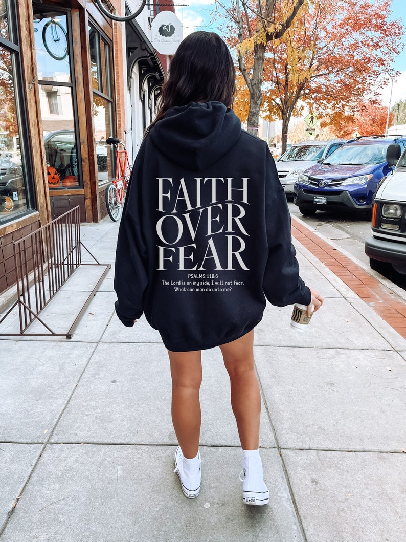 Op de afbeelding: Een zwarte hoodie met de woorden "FAITH OVER FEAR" gedrukt op de achterkant. De hoodie bevat ook een Bijbelvers uit Psalm 118:6, "De HEERE is mijn helper, ik zal niet vrezen; wat kan de mens mij doen?"