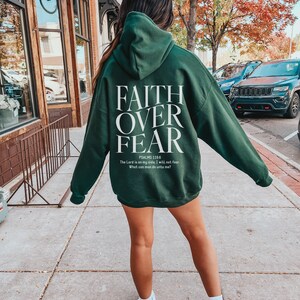 Op de afbeelding: Een donkergroen hoodie met de woorden "FAITH OVER FEAR" in wit gedrukt op de achterkant. De tekst "PSALMS 118.6" is onder de woorden gedrukt, gevolgd door de quote "The Lord is on my side, I will not fear. What can man do unto me?"