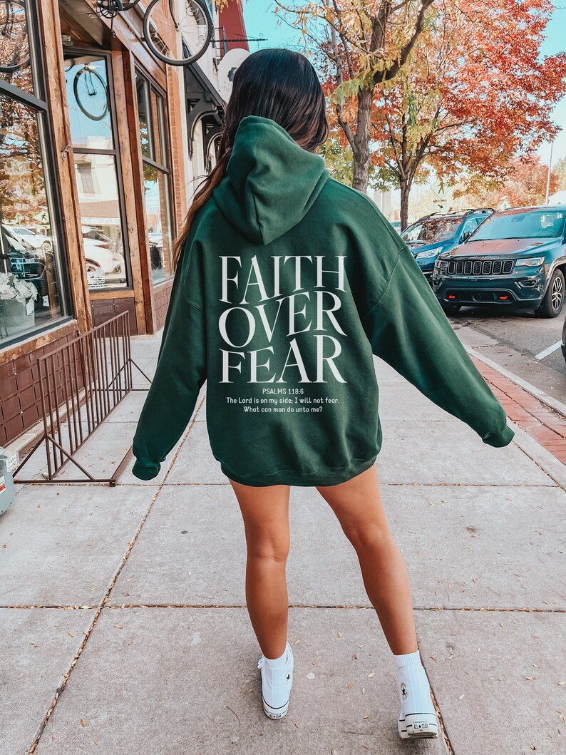 Op de afbeelding: Een donkergroen hoodie met de woorden "FAITH OVER FEAR" in wit gedrukt op de achterkant. De tekst "PSALMS 118.6" is onder de woorden gedrukt, gevolgd door de quote "The Lord is on my side, I will not fear. What can man do unto me?"