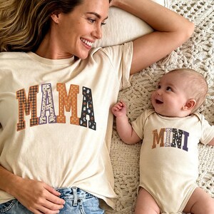 May include: A cream-colored short-sleeve t-shirt with the word MAMA in faux patchwork letters and a matching cream-colored baby bodysuit with the word MINI in faux patchwork letters. The letters feature various patterns including plaid and polka dots.
