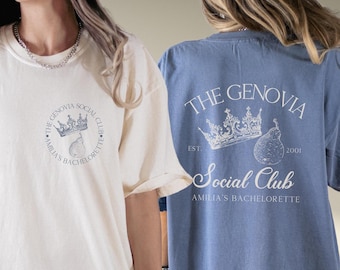 May include: Two t-shirts with a graphic design featuring a crown, a pear, and the text "The Genovia Social Club, Amilia's Bachelorette, Est. 2001". The t-shirt on the left is ivory and the t-shirt on the right is blue jean.