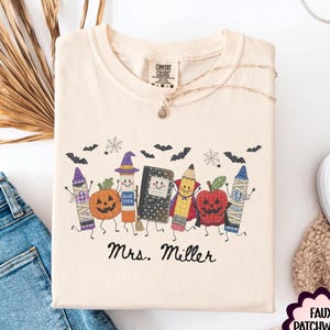 May include: A cream-colored t-shirt featuring a graphic print of anthropomorphic school supplies including a pumpkin, glue stick, notebook, pencil, and apple, all with faces. The text Mrs. Miller is printed below the characters with bats and webs.