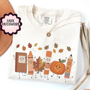 May include: An ivory t-shirt featuring a graphic print of a notebook, crayon, leaf, glue stick, pumpkin, and pencil with smiling faces. The design includes small scattered autumn leaves and the text Faux Patchwork. The shirt has a Comfort Colors label.