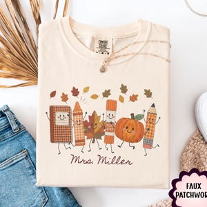May include: A cream-colored t-shirt featuring a graphic of a smiling notebook, crayon, leaf, glue stick, pumpkin, and pencil with arms and legs. The text "Mrs. Miller" is printed below the characters, surrounded by falling autumn leaves.