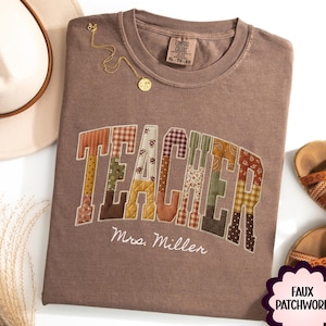 May include: A brown short-sleeved t-shirt featuring the word TEACHER in a faux patchwork design with various floral and plaid patterns. Below the word, the name Mrs. Miller is printed in white script. The shirt has a crew neckline and a tag.