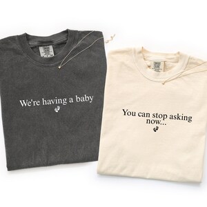 May include: A dark gray t-shirt with the white text We're having a baby and a small footprint icon, and a cream t-shirt with the black text You can stop asking now... and a small footprint icon. Both shirts feature Comfort Colors brand labels.