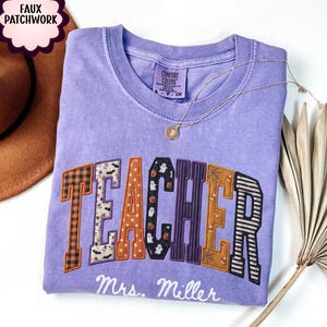 May include: A light purple short-sleeve t-shirt featuring the word TEACHER in large, faux patchwork letters with various Halloween patterns including ghosts, pumpkins, and spiderwebs. Below the word, the text Mrs. Miller is printed in white.