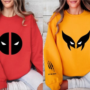 May include: Two sweatshirts, one red and one yellow, with black superhero logos. The red sweatshirt features a Deadpool logo, while the yellow sweatshirt has a Wolverine logo. Both sweatshirts have a date printed on the sleeve.