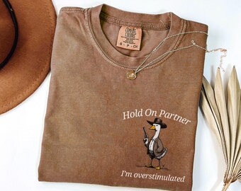 May include: A brown short-sleeve t-shirt featuring a graphic of a duck wearing a cowboy hat and holding a small gun. The text above the duck reads Hold On Partner and the text below reads I'm overstimulated. The shirt has a crew neckline.