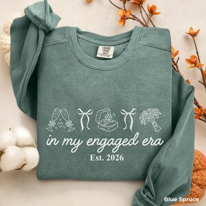 Blue Spruce Comfort Colors sweatshirt with white In My Engaged Era Est. 2026 design, champagne glasses, bows, engagement ring and bridal bouquet. Cozy bride to be crewneck for newly engaged women and engagement gifts.