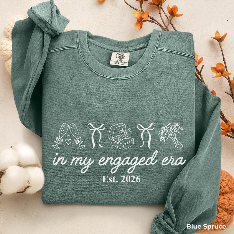 Blue Spruce Comfort Colors sweatshirt with white In My Engaged Era Est. 2026 design, champagne glasses, bows, engagement ring and bridal bouquet. Cozy bride to be crewneck for newly engaged women and engagement gifts.
