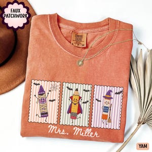 May include: A burnt orange crewneck t-shirt featuring a graphic of three postage stamp-style illustrations. The designs show a mummy crayon, a vampire pencil, and a witch glue stick. The text Mrs. Miller is printed below the illustrations.