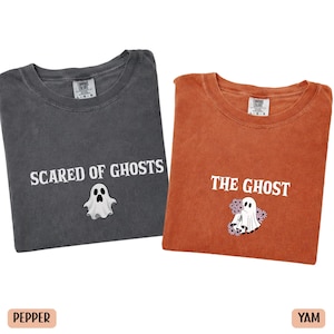 May include: A charcoal grey t-shirt with the white text SCARED OF GHOSTS and a ghost graphic, alongside a burnt orange t-shirt with the white text THE GHOST and a ghost graphic surrounded by small purple flowers, both featuring crew necklines.