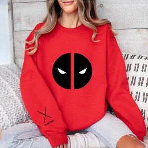 May include: Red sweatshirt with a black Deadpool logo and the text "07-28-2024" and a pair of crossed swords on the sleeve.