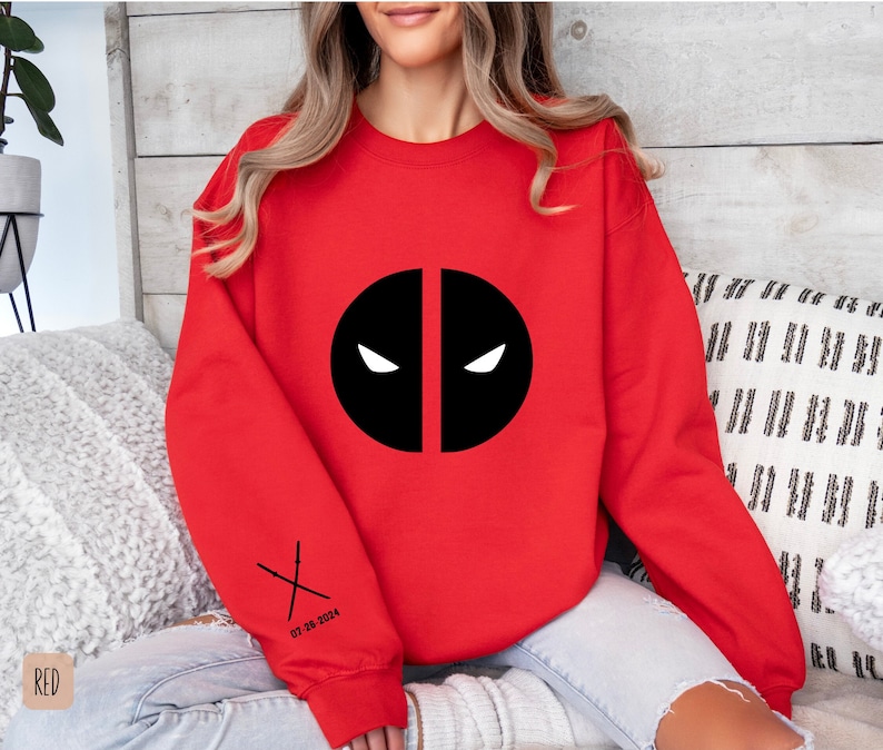 May include: Red sweatshirt with a black Deadpool logo and the text "07-28-2024" and a pair of crossed swords on the sleeve.