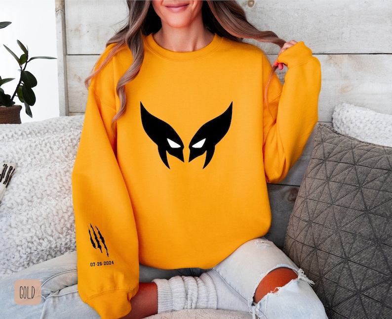 May include: A yellow sweatshirt with a black Wolverine mask graphic on the front and black claw marks on the left sleeve. The text "07.26.2024" is printed on the sleeve.