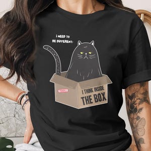 Könnte beinhalten: Schwarzes T-Shirt mit einer Cartoon-Katze in einem Pappkarton. Die Katze hat einen mürrischen Gesichtsausdruck, mit dem Text "I THINK INSIDE THE BOX" unter der Box. Über der Katze steht der Text "I NEED TO BE DIFFERENT."
