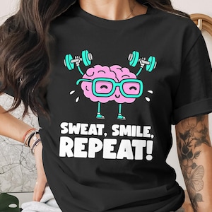 Könnte beinhalten: Schwarzes T-Shirt mit einem Cartoon-Gehirn, das Gewichte hebt, eine Brille trägt und lächelt. Der Text "SWEAT, SMILE, REPEAT!" ist in Weiß unter der Grafik aufgedruckt. Ein lustiges und motivierendes Design für Fitness-Enthusiasten.